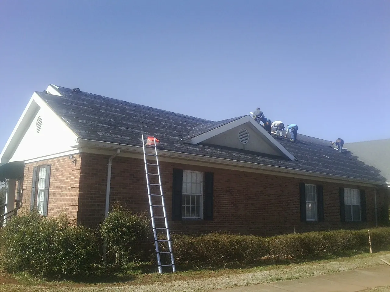 Crew tearing shingles off a brick ranch roof with a ladder set — Metal Roof Repair work by our crew in Balsam Grove