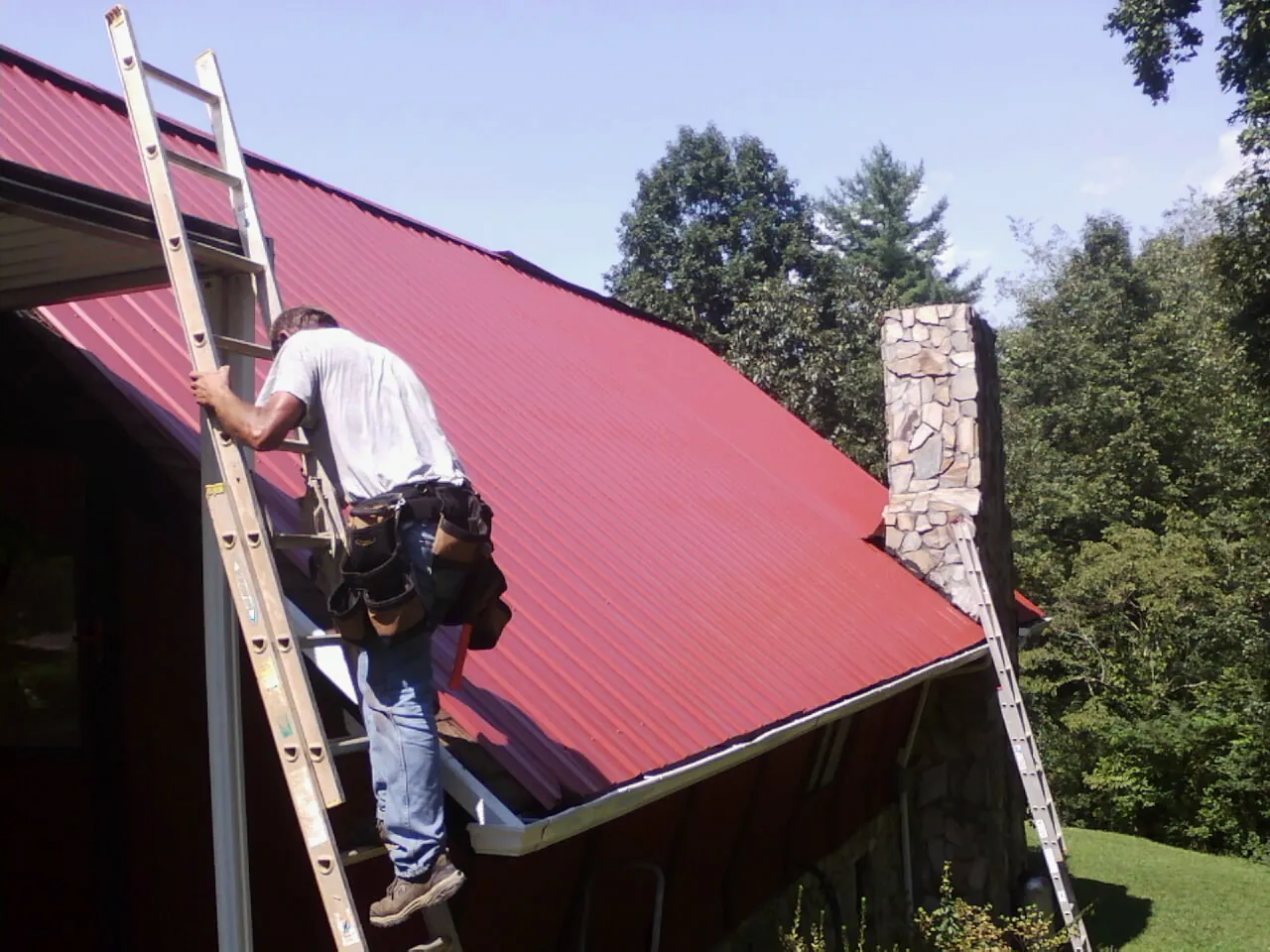 Roofer on a ladder beside a red metal roof and stone chimney — Soffit & Fascia Repair by Roofing Brothers in Balsam Grove